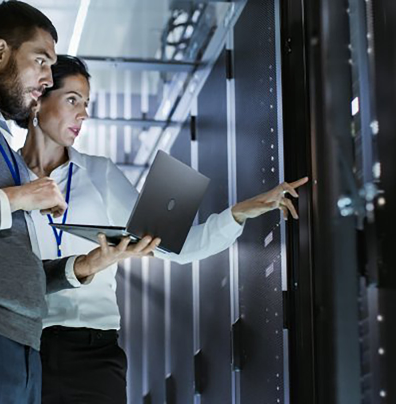 A man holding a laptop while a woman points at a panel in a server room