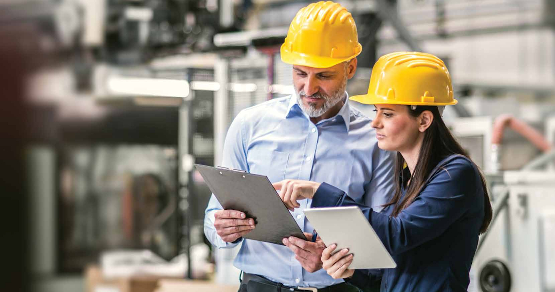 Two people in safety equipment looking at a clipboard with a blurred factory background